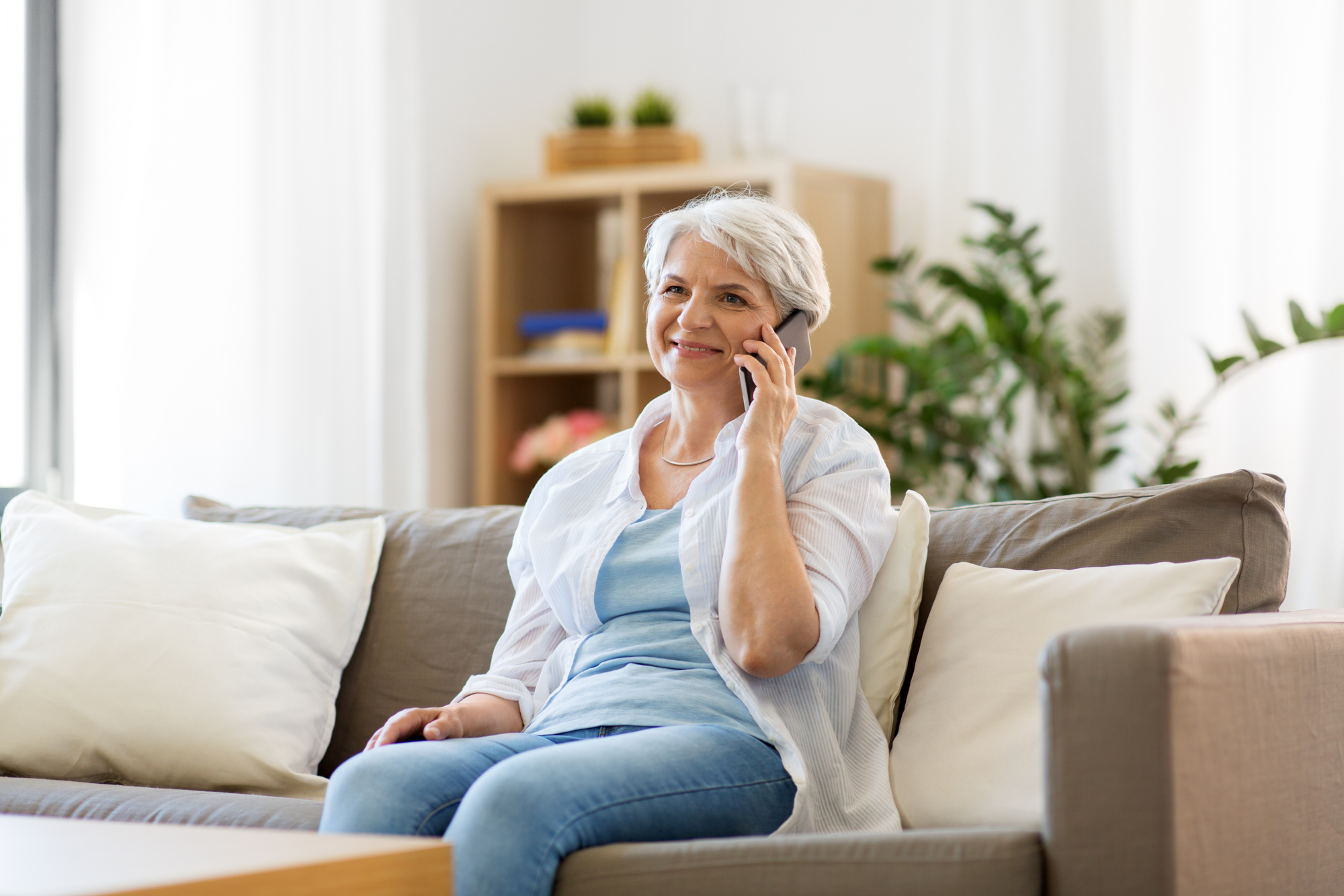 Smiling older woman talking on a smartphone while sitting comfortably on a couch in a bright, airy living room.