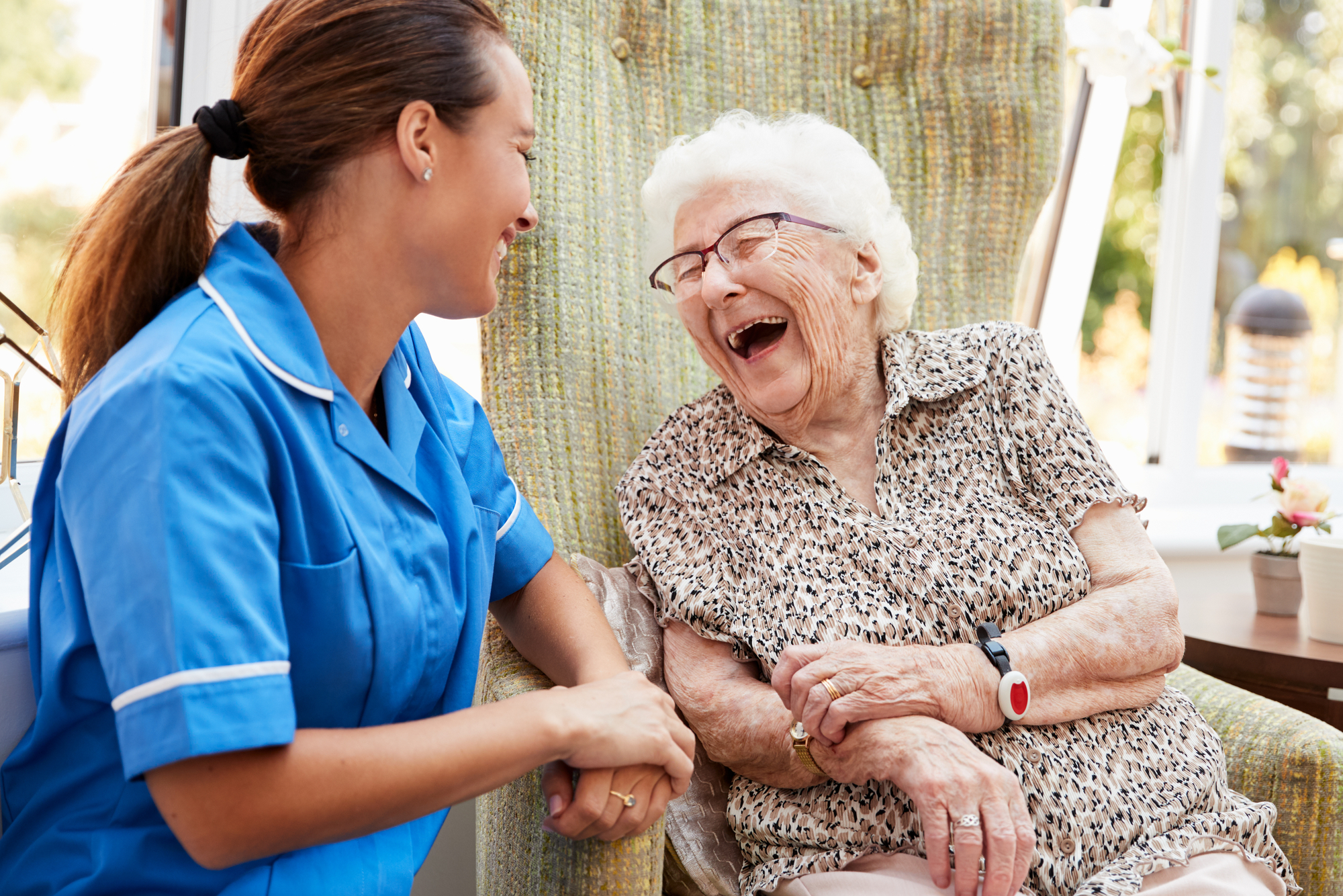 A caregiver in a blue uniform shares a joyful moment with a laughing elderly woman, highlighting the importance of companionship in elder care.
