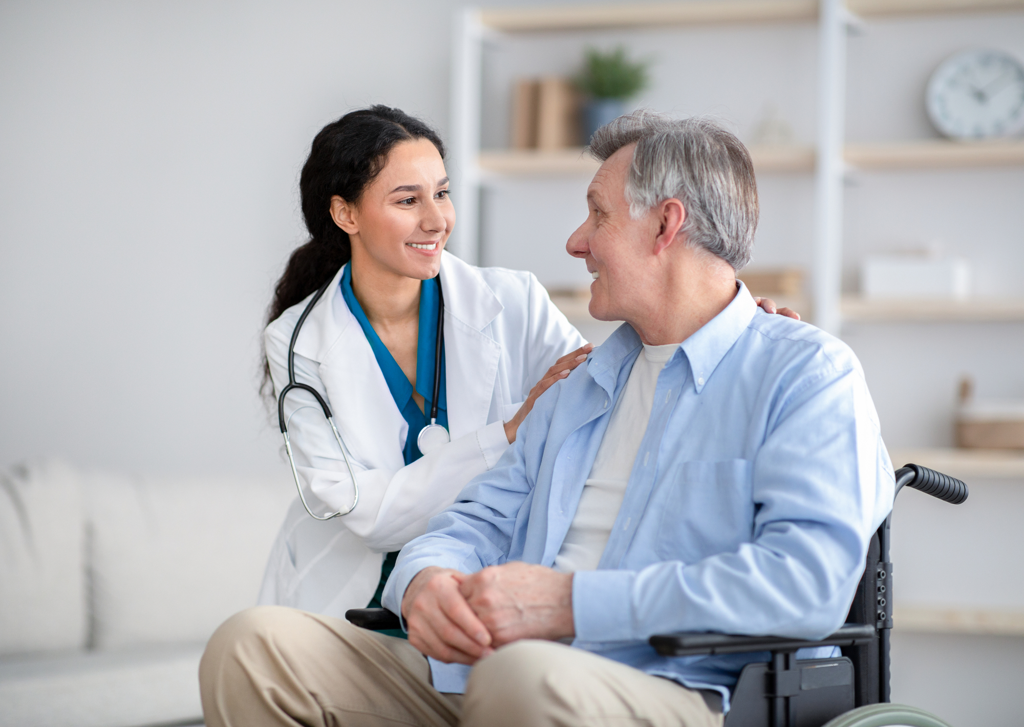 A caregiver, wearing a blue uniform, smiles and gently touches the shoulder of an elderly man in a gray cardigan, fostering a warm, supportive moment.