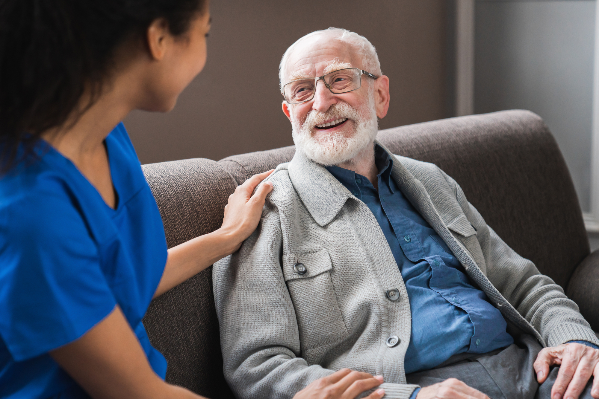 A caregiver, wearing a blue uniform, smiles and gently touches the shoulder of an elderly man in a gray cardigan, fostering a warm, supportive moment.