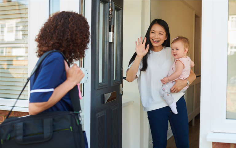 A smiling mother waves at a healthcare worker standing at her doorstep, holding her baby in her arms, indicating a home visit.
