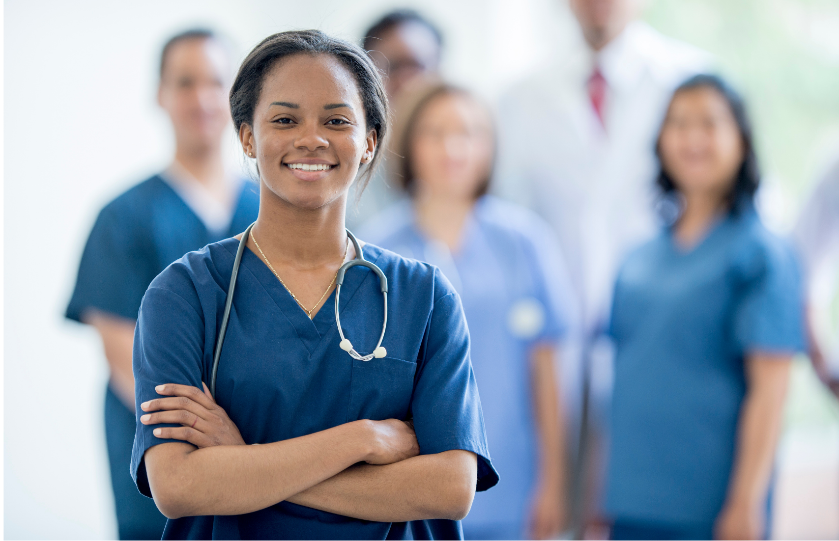 A confident nurse in scrubs stands in front of a blurred team of healthcare professionals, highlighting teamwork and dedication in healthcare.