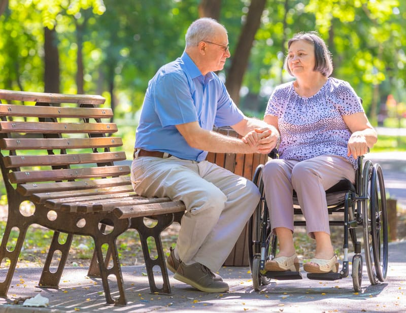 An elderly man and woman share a tender moment in a park. He holds her hands while she sits in a wheelchair, surrounded by greenery.